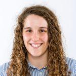 A young woman with long, curly brown hair smiles at the camera. She is wearing a blue and white striped shirt and is posed against a plain white background.