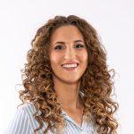 A woman with long, curly light brown hair smiles at the camera. She is wearing a light blue and white striped shirt and is posed in front of a plain white background.